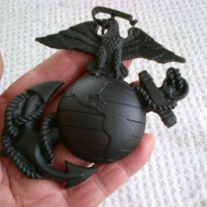May include: A black metal eagle, globe, and anchor emblem. The eagle is perched on top of the globe, with its wings spread out. The anchor is below the globe, with a rope wrapped around it.