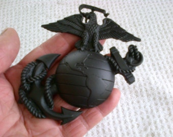 May include: A black metal eagle, globe, and anchor emblem. The eagle is perched on top of the globe, with its wings spread out. The anchor is below the globe, with a rope wrapped around it.