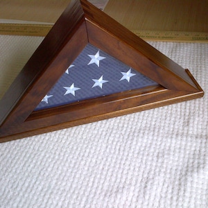 May include: A triangular wooden display case with a dark brown finish, containing a folded American flag with visible white stars on a blue field. The case is positioned next to a yellow ruler showing measurements in inches.