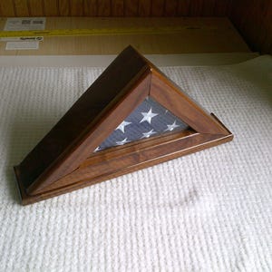 May include: A wooden flag display case in a triangular shape. The case is made of dark brown wood and features a blue background with white stars. The case is designed to hold a folded American flag.