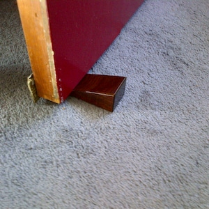 May include: A dark wood doorstop wedges a door open. The doorstop is a dark brown, rectangular prism. The door is red with a wooden frame.