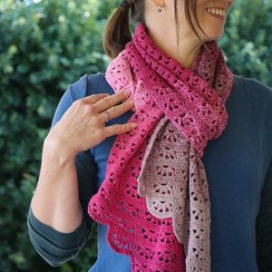 Puede incluir: Una bufanda de ganchillo rosa y marrón con un patrón de encaje. La bufanda está drapeada alrededor del cuello de una persona que lleva una camisa azul.