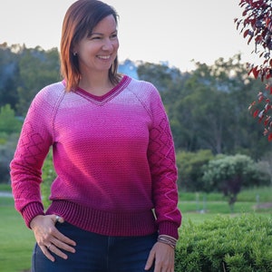 Può includere: Un maglione a ombre con sfumature rosa e bordeaux, con scollo a V e un motivo a treccia sulle maniche.