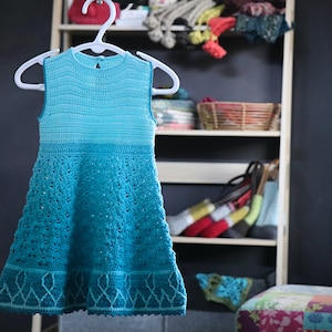 Könnte beinhalten: Ein ärmelloses Häkelkleid mit einem blauen und türkisfarbenen Ombre-Muster. Das Kleid hat einen gewellten Saum und ein zartes Spitzenmuster.