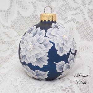 May include: A spherical dark blue Christmas ornament featuring hand-painted white floral designs with layered petals and small iridescent accents at the centers. The ornament has a gold-colored metal cap with a silver hanging loop and the text Margot Clark.