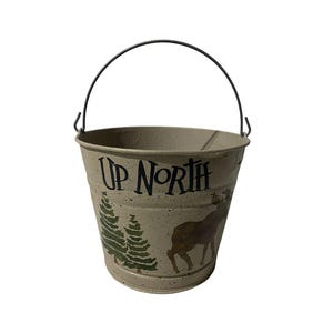 May include: Tan metal bucket with a black handle. The bucket has the words "UP NORTH" printed on the front, along with a green tree and a brown deer illustration. The bucket is designed for decorative or functional use.