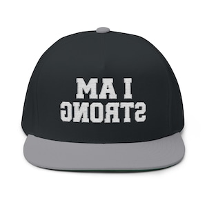 May include: A black snapback cap with a grey brim and a grey button on top. The front features white embroidered text that reads I AM STRONG, with the word STRONG printed in an inverted, upside-down orientation relative to the top line.
