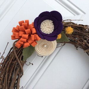 May include: A handmade grapevine wreath decorated with felt flowers in shades of orange, purple, and cream. The wreath features a cluster of orange felt loops, a purple flower with a cream center, and a cream-colored spiral flower.