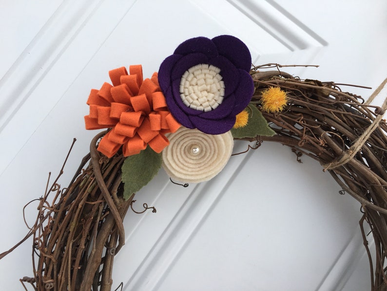May include: A handmade grapevine wreath decorated with felt flowers in shades of orange, purple, and cream. The wreath features a cluster of orange felt loops, a purple flower with a cream center, and a cream-colored spiral flower.