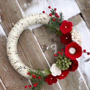 May include: A white birch wreath decorated with red felt flowers, green felt leaves, and sprigs of red berries.