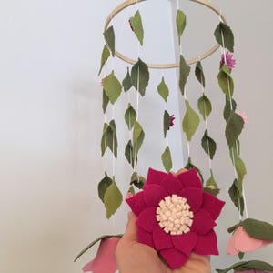 May include: A wooden ring mobile with felt leaves and flowers in shades of pink and green. The mobile is hanging from the ceiling.