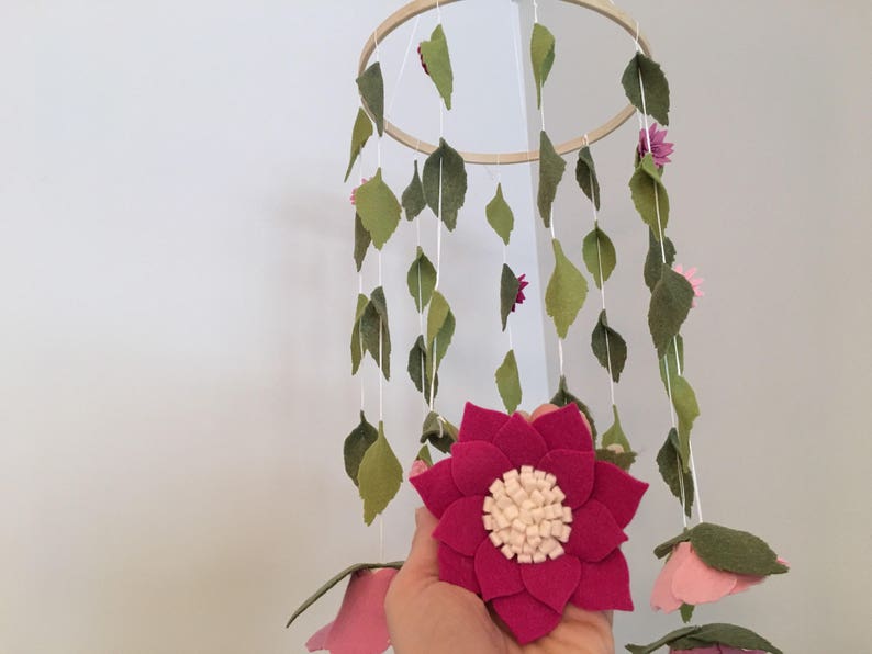 May include: A wooden ring mobile with felt leaves and flowers in shades of pink and green. The mobile is hanging from the ceiling.