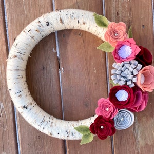 May include: A white birch wreath decorated with felt flowers in shades of pink, red, and grey. The flowers are clustered on the right side of the wreath.