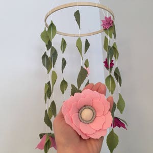 May include: A pink felt flower with a white center hangs from a wooden ring with green felt leaves. The mobile is a nursery decoration.