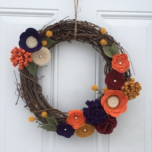 May include: A decorative wreath made of intertwined brown twigs, adorned with felt flowers in shades of orange, purple, cream, and burgundy. Green felt leaves and small yellow accents add to the autumnal theme. The wreath is suspended by a natural twine.