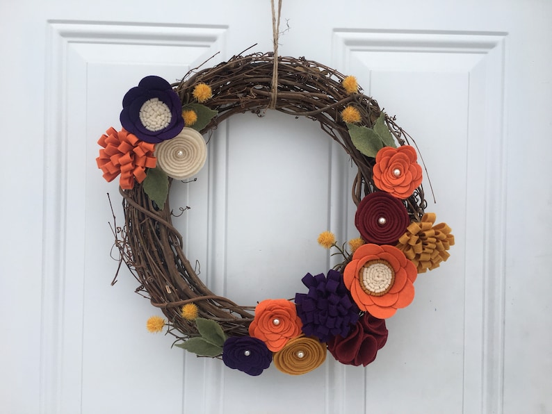 May include: A decorative wreath made of intertwined brown twigs, adorned with felt flowers in shades of orange, purple, cream, and burgundy. Green felt leaves and small yellow accents add to the autumnal theme. The wreath is suspended by a natural twine.