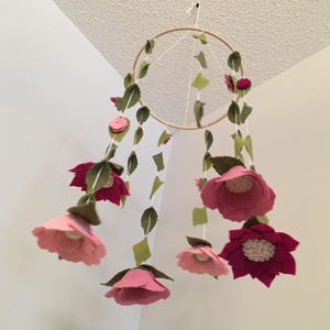 May include: A pink and green felt flower mobile hanging from a wooden hoop. The mobile features multiple pink flowers with green leaves and a white center.