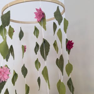 May include: A wooden hoop mobile with felt leaves and flowers in shades of pink and green. The mobile is hanging from the ceiling.