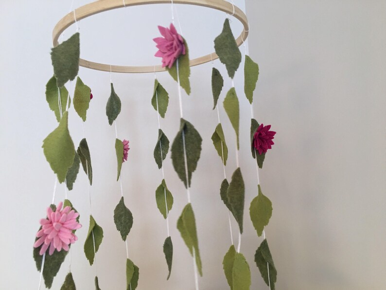 May include: A wooden hoop mobile with felt leaves and flowers in shades of pink and green. The mobile is hanging from the ceiling.