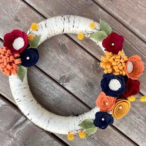 Peut inclure: Une couronne en écorce de bouleau décorée de fleurs en feutre dans des tons de rouge, orange, jaune et bleu. Les fleurs sont disposées en cercle autour de la couronne.