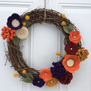 May include: A grapevine wreath adorned with colorful felt flowers in shades of orange, purple, burgundy, and cream. The wreath features green felt leaves and small yellow pom-poms, creating a vibrant and seasonal decorative piece.
