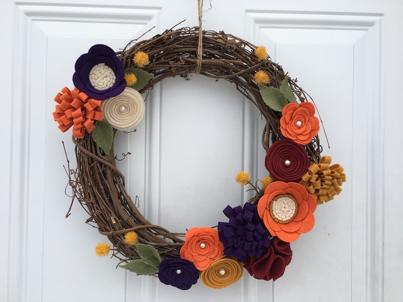 May include: A grapevine wreath adorned with colorful felt flowers in shades of orange, purple, burgundy, and cream. The wreath features green felt leaves and small yellow pom-poms, creating a vibrant and seasonal decorative piece.