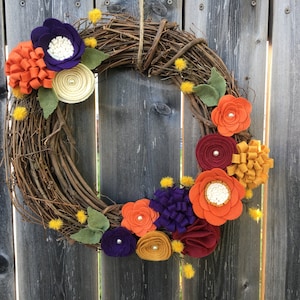 May include: A handmade wreath with a grapevine base, adorned with colorful felt flowers in shades of orange, purple, burgundy, and yellow. The wreath is decorated with small yellow pom-poms and green felt leaves. A decorative piece for the front door.