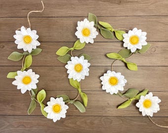 May include: A felt garland with white daisies and green leaves. The garland is strung together with twine.