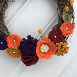 May include: A handcrafted grapevine wreath adorned with colorful felt flowers. The wreath features orange, purple, burgundy, and yellow flowers with pearl accents and green leaves. A decorative piece for the home.