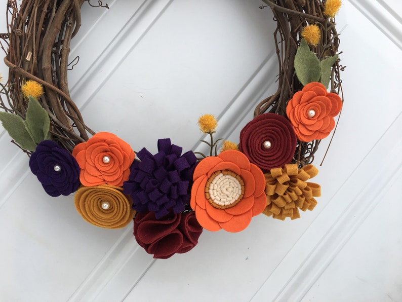 May include: A handcrafted grapevine wreath adorned with colorful felt flowers. The wreath features orange, purple, burgundy, and yellow flowers with pearl accents and green leaves. A decorative piece for the home.