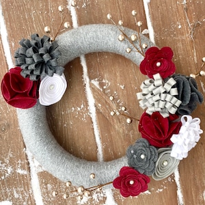 Peut inclure: Une couronne faite à la main avec une base en feutre gris, ornée de fleurs en feutre dans les tons de rouge, de gris et de blanc. La couronne est décorée de brins de baies dorées, créant une pièce festive et décorative pour la maison.