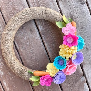 May include: A burlap wreath decorated with felt flowers in shades of pink, yellow, blue, and purple. The flowers are arranged in a cluster on the right side of the wreath.