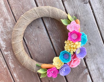 May include: A burlap wreath decorated with felt flowers in shades of pink, yellow, blue, and purple. The flowers are arranged in a cluster on the right side of the wreath.