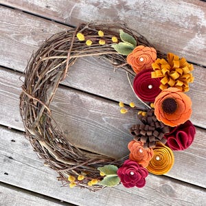 Peut inclure: Une couronne d'automne en brindilles ornée de fleurs en feutre dans des tons de rouge, orange et jaune. La couronne est décorée de petites boules de feutre jaunes.