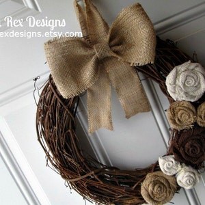 May include: A brown twig wreath with a burlap bow and brown, white, and beige fabric flowers. The wreath is hanging on a white door.