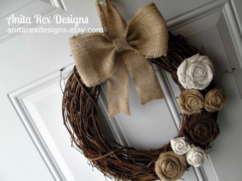May include: A brown twig wreath with a burlap bow and brown, white, and beige fabric flowers. The wreath is hanging on a white door.