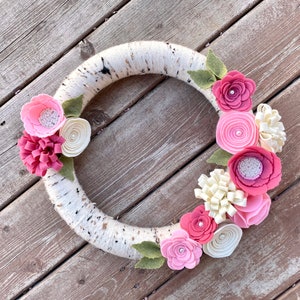May include: A birch bark wreath decorated with pink and white felt flowers. The wreath is adorned with leaves and has a rustic, natural look.