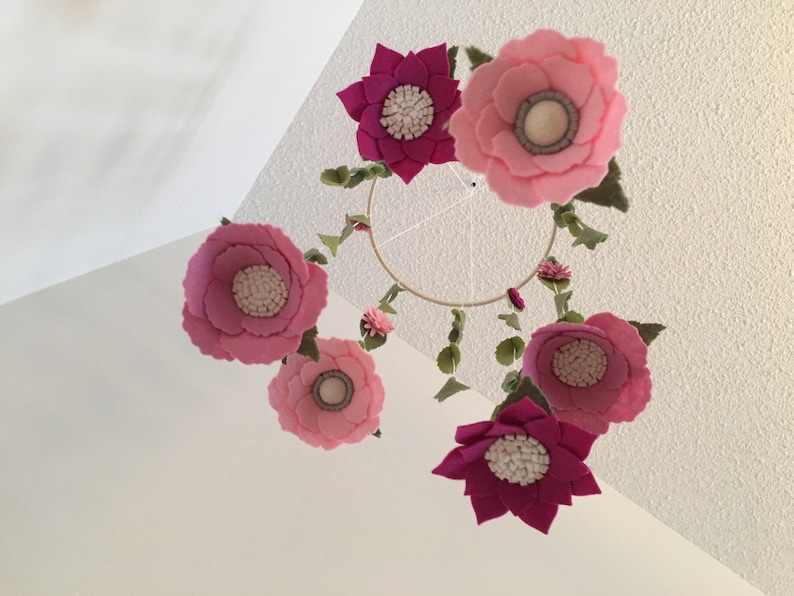 May include: A pink and magenta felt flower mobile with a circular metal frame. The mobile features six flowers with white centers and green felt leaves.