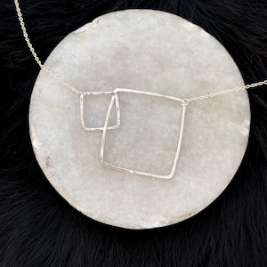 Puede incluir: Un collar plateado con dos colgantes cuadrados superpuestos. Los colgantes tienen una textura martillada y están suspendidos de una delicada cadena. El collar se muestra sobre una piedra circular gris claro sobre un fondo negro.