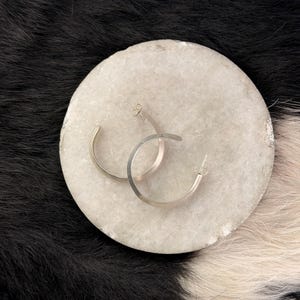 Puede incluir: Un par de pendientes de aro plateados sobre una piedra redonda y clara. Los pendientes tienen un diseño sencillo y moderno, con un acabado metálico liso. El fondo presenta una superficie con estampado en blanco y negro.