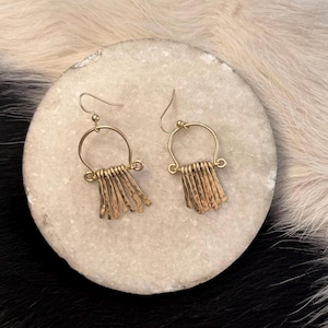 Puede incluir: Un par de pendientes dorados. Cada pendiente presenta una parte superior circular con una serie de barras verticales texturizadas colgando debajo. Los pendientes se exhiben sobre una piedra redonda de color claro sobre un fondo blanco y negro.