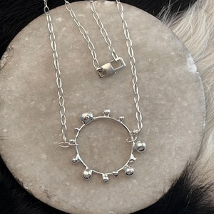 May include: A silver-toned necklace featuring a circular pendant with irregular, textured embellishments. The pendant is suspended from a delicate chain with a simple clasp. The necklace rests on a light gray, textured surface.