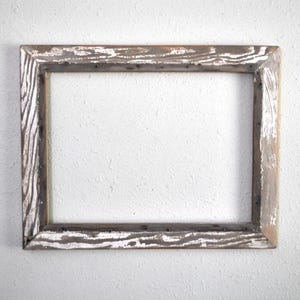 May include: A rectangular wooden picture frame with a distressed, rustic appearance. The frame is made of light brown wood with white paint accents, and it is empty. The frame is mounted on a white wall.