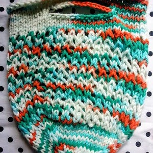 May include: Hand-knitted bag in a colorful pattern. The bag features a mix of teal, orange, and white yarns, creating a textured design. The top has a handle for carrying. The bag is made of a soft, flexible material.