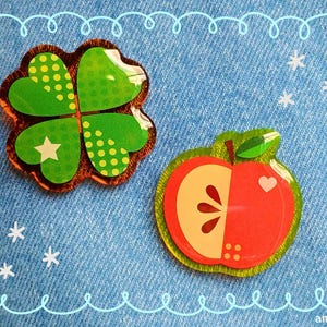 Pode incluir: Dois broches decorativos sobre um fundo de ganga azul. Um é um trevo de quatro folhas com detalhes verdes e amarelos. O outro é uma maçã vermelha com um coração e uma folha verde. Acentos brancos de estrelas e flocos de neve.