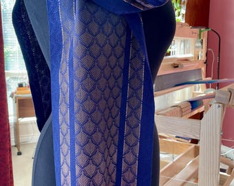 May include: A navy blue scarf with a geometric pattern in shades of gold and blue. The scarf is draped over a mannequin, showcasing its design and fringe detail. The scarf's design features a repeating leaf-like pattern.