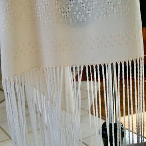 May include: White woven shawl with a fringe border. The shawl is made of a fine, delicate fabric and has a subtle pattern.