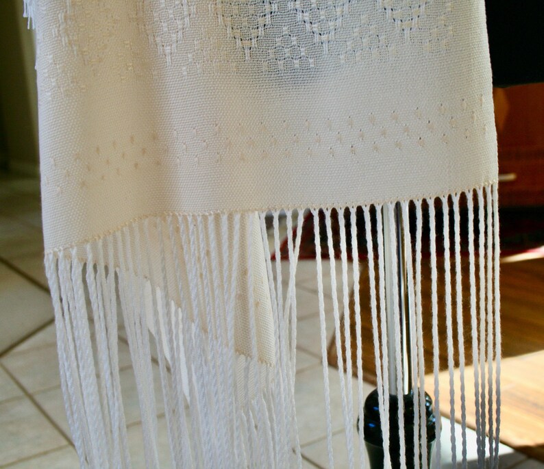 May include: White woven shawl with a fringe border. The shawl is made of a fine, delicate fabric and has a subtle pattern.