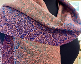 May include: A woven scarf featuring a complex geometric pattern in contrasting shades of coral pink and deep royal blue. The fabric displays a dense, intricate texture with repeating diamond-like motifs throughout, draped over a black mannequin form.