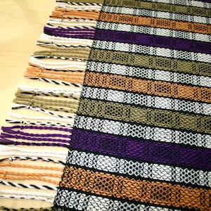 May include: A woven tapestry with a fringe border. The tapestry features a striped pattern in shades of orange, purple, green, white, and black.